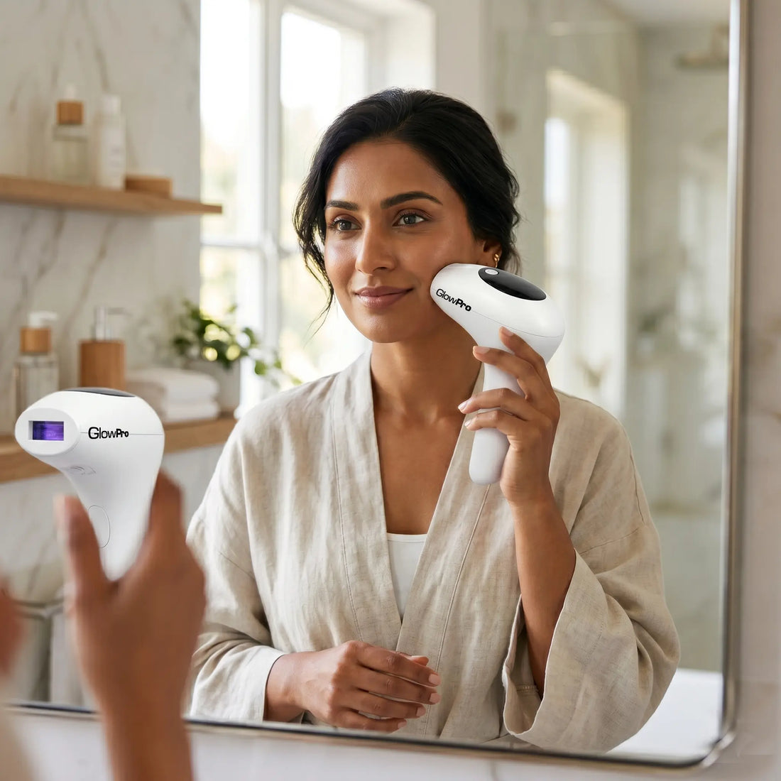 A Pakistani woman wearing a light linen robe standing in front of a bathroom mirror, applying a GlowPro IPL hair removal device to her face, with the mirror reflecting the front of the handset.
